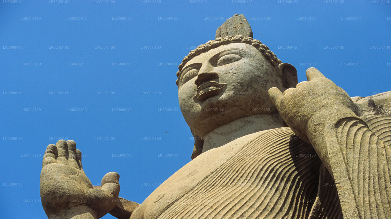 Stone Buddha statue with one hand raised in blessing gesture against a clear blue sky.