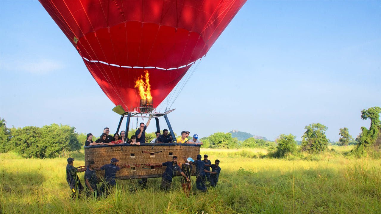 A hot air balloon is inflated and ready for flight with people gathered around, set against a scenic landscape.